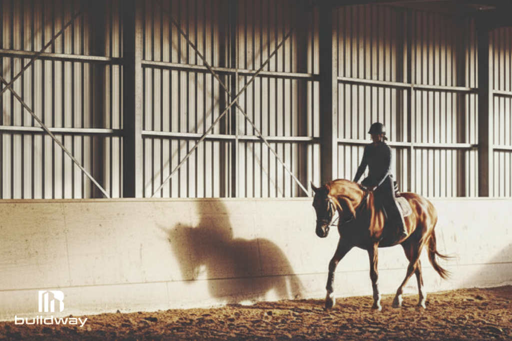 Rider training a horse inside an indoor agricultural riding arena with steel walls and soft dirt flooring, designed by Buildway.
