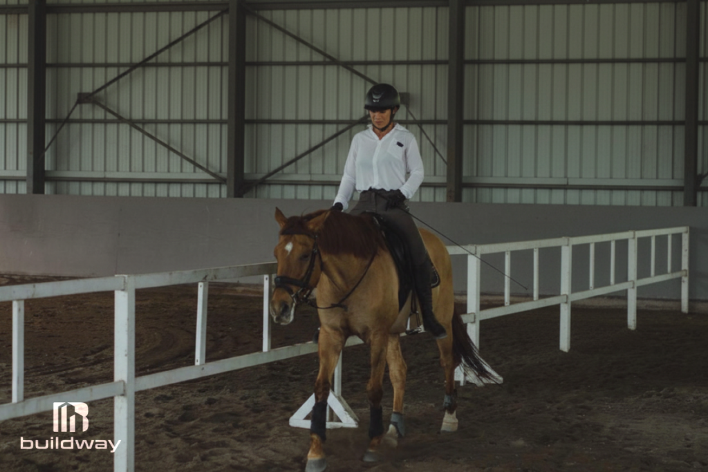 Rider in a white shirt practicing horseback riding inside an enclosed agricultural riding arena with steel walls and fenced training lanes, designed by Buildway.