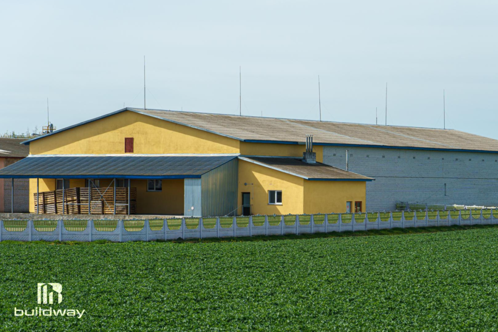 Yellow and gray agricultural storage building with a fenced perimeter, situated beside a green crop field, designed by Buildway.