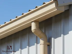 Beige steel building gutter and downspout system installed along the roof edge of a Buildway I-beam structure.