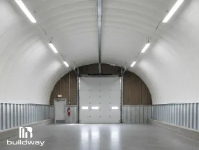 Interior view of a Quonset-style steel building by Buildway featuring bright insulated walls and ceiling, a large roll-up door, and modern lighting fixtures.