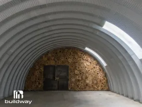 Interior view of a Quonset prefab steel building by Buildway, showing the curved corrugated metal structure with a stacked wood wall at the end and concrete flooring.