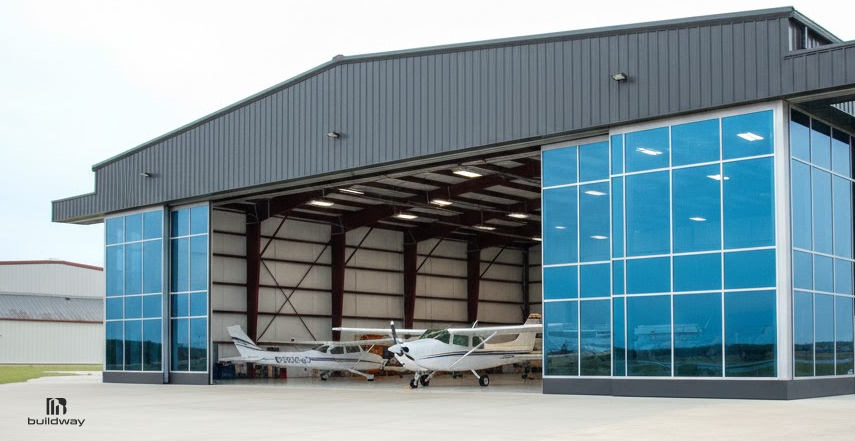 How Long Do Steel Buildings Last If You Ignore Maintenance? The Result Is Brutal 2 Large steel aircraft hangar with wide open doors, blue-tinted glass panels, and small airplanes parked inside on a concrete apron.
