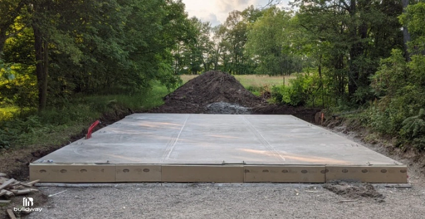 Freshly poured concrete slab foundation in a clearing surrounded by trees, prepared for a future steel building installation.