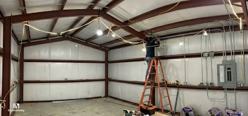 How to Personalize a Steel Building Without Wasting Money on the Wrong Parts 9 Person installing electrical wiring inside an insulated steel building with exposed red framing, conduit, and a visible breaker panel.