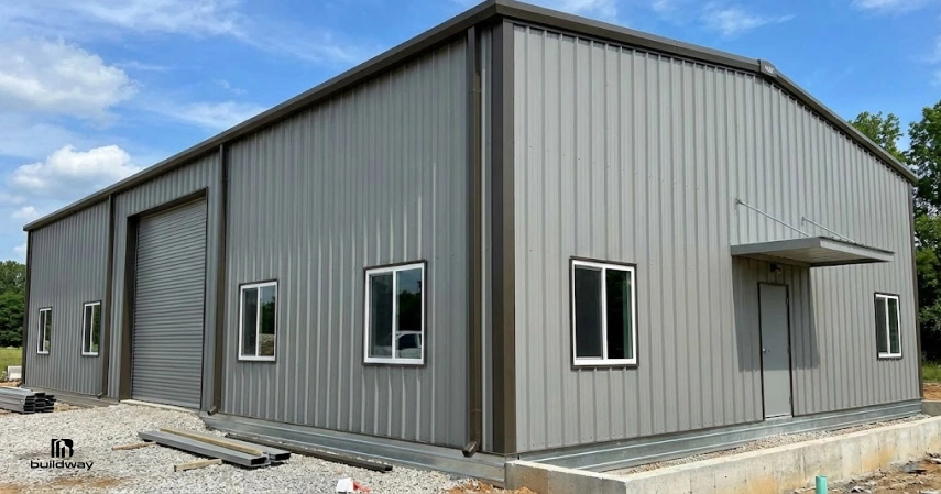 Steel Building Prices in 2026: What You’ll Really Pay 6 Completed steel building with windows, a roll-up door, and a side entrance, shown on a gravel lot.